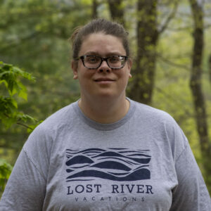 A person with glasses is standing outdoors in front of a blurred forest background. They are wearing a gray T-shirt with a "Lost River Vacations" logo on it. The person has their hair pulled back and is smiling slightly.